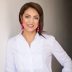 Amalia Sterescu headshot photo, smiling woman wearing pink earrings and white top leaning against a white wall