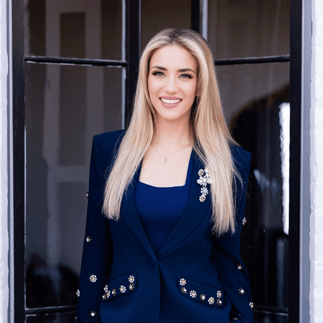 Casey Hughes headshot photo, smiling blond woman wearing navy blazer and dress in front of window