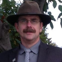 James Greengrass PCC headshot photo, smiling man with hat and glasses wearing a blazer collared shirt and tie outside