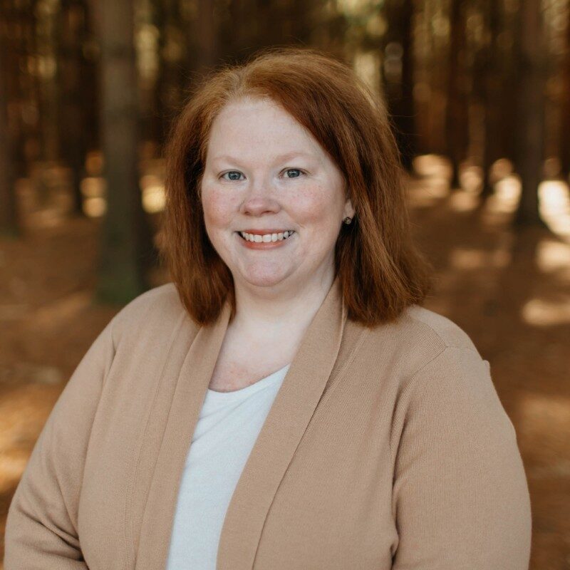 Headshot of a woman standing in front of a forest of trees.