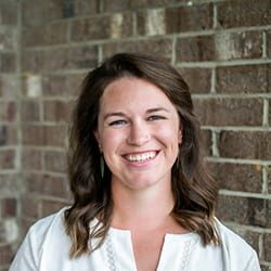 Laura Shrake headshot photo, smiling woman with brown hair and a white top standing in front of a brick wall