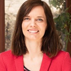 Lenka Grackova ACC PCC headshot photo, smiling woman with brown hair and red blazer in front of greenery