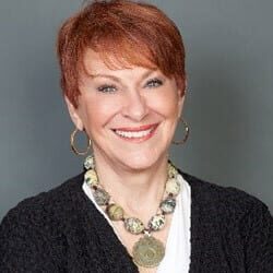 Valerie Sokolosky PCC headshot photo, smiling woman with short red hair wearing a dark top earrings and a necklace in front of a gray background