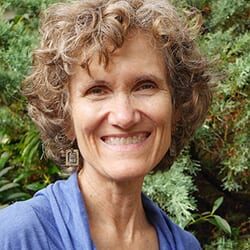 Dee Wagner headshot photo, woman smiling with curly brown hair and blue top in front of greenery