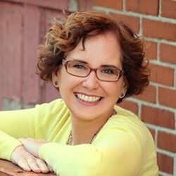 Aimee Bucher ACC PCC headshot photo, smiling woman with brown hair wearing yellow top and red glasses in front of a brick wall
