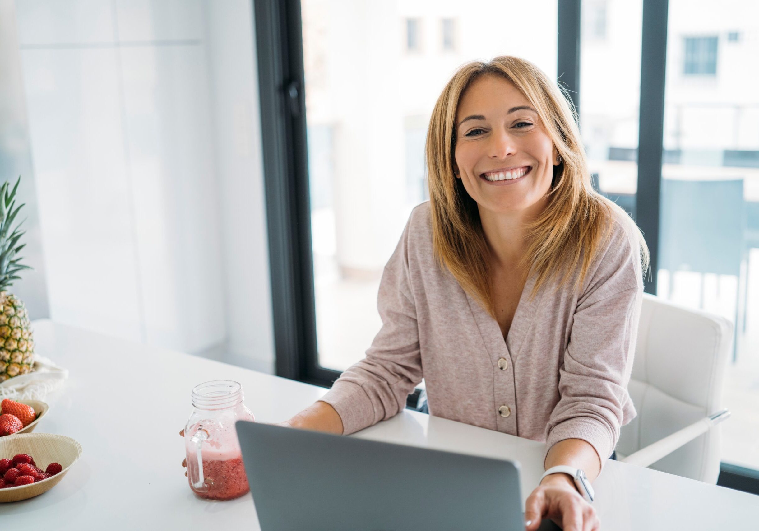 Smiling,Woman,Holding,Smoothie,By,Laptop,In,Bright,Kitchen