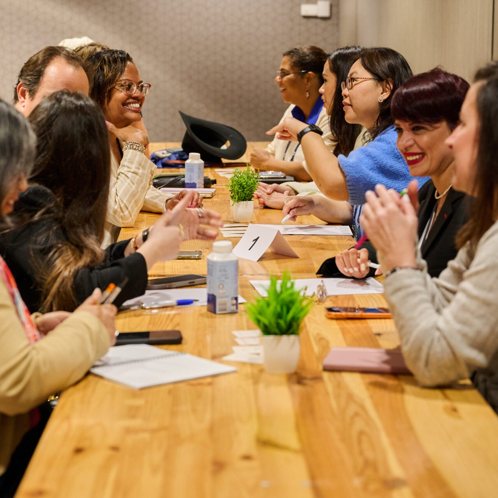 A diverse group of men and women seated at a long community table, seen having a discussion and smiling.