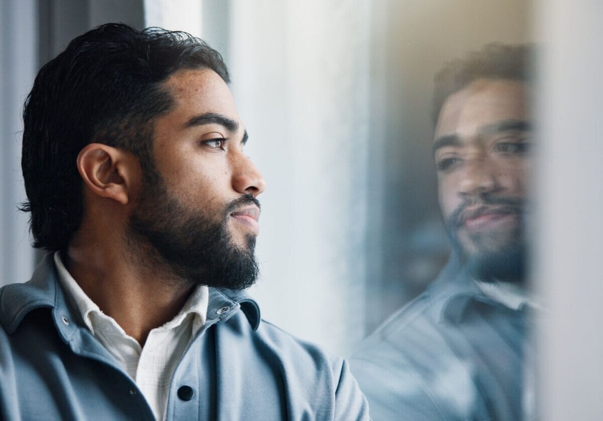 Man gazing thoughtfully through a window with his reflection visible, symbolizing coaching self-awareness