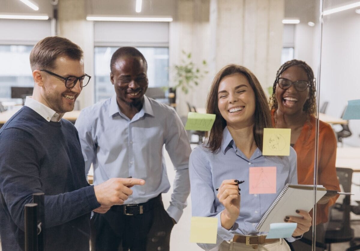 Four coworkers smiling and collaborating around a glass board covered with colorful sticky notes in a bright office.