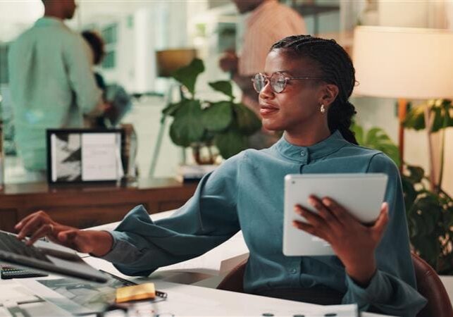 Professional Black woman in teal blouse working at desk with laptop and tablet in modern office with plants and colleagues.