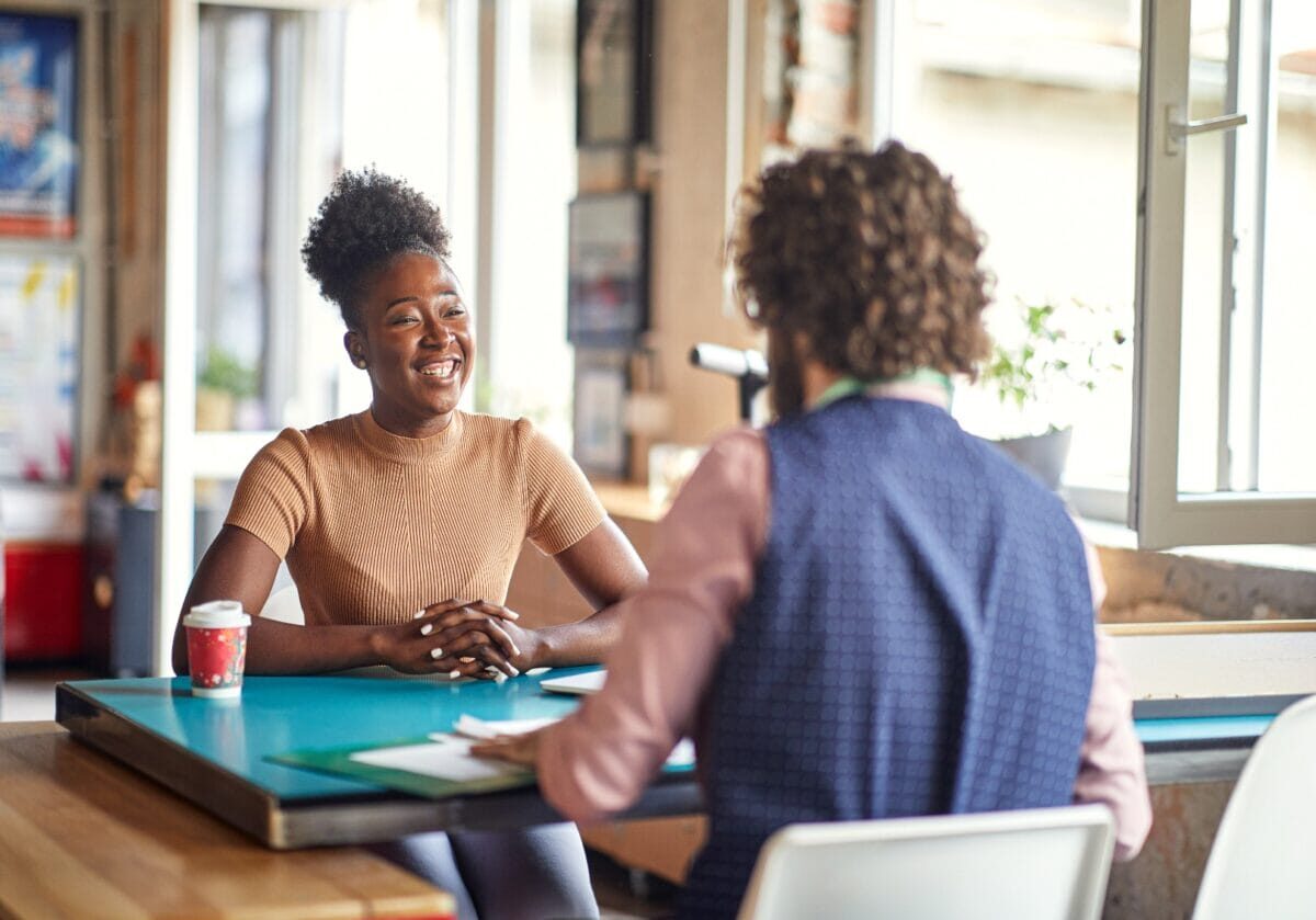 A smiling woman with her hair in a high bun sits at a teal desk during an interview or meeting with a man whose back is to the camera.