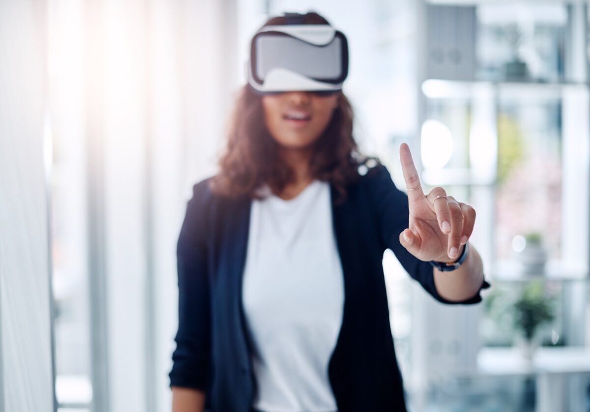A woman wearing a VR headset points her finger forward to interact with a virtual interface in a bright office setting.