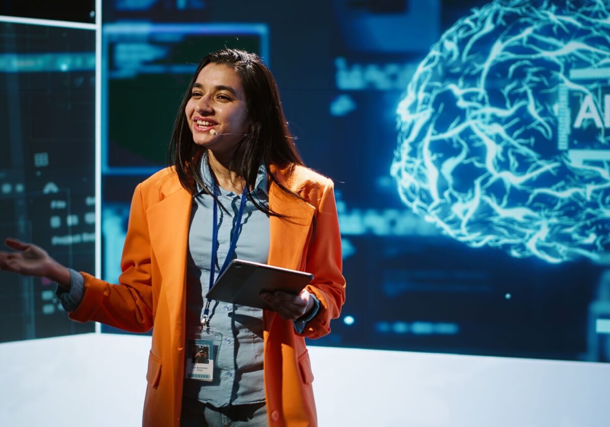A smiling female speaker in a bright orange blazer holds a tablet and gestures during an AI technology presentation.
