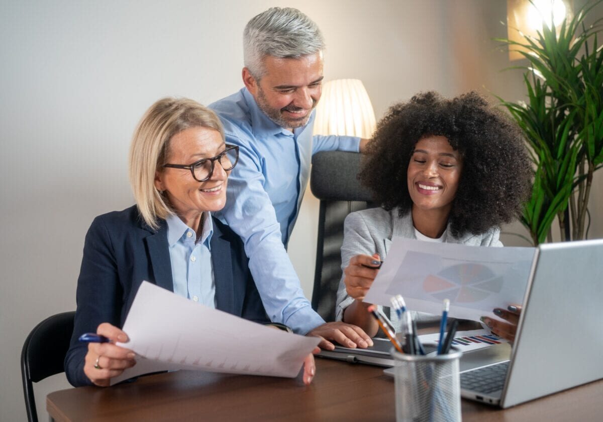 Three business professionals smile while reviewing printed data reports and charts together at a desk with a laptop during an office meeting.