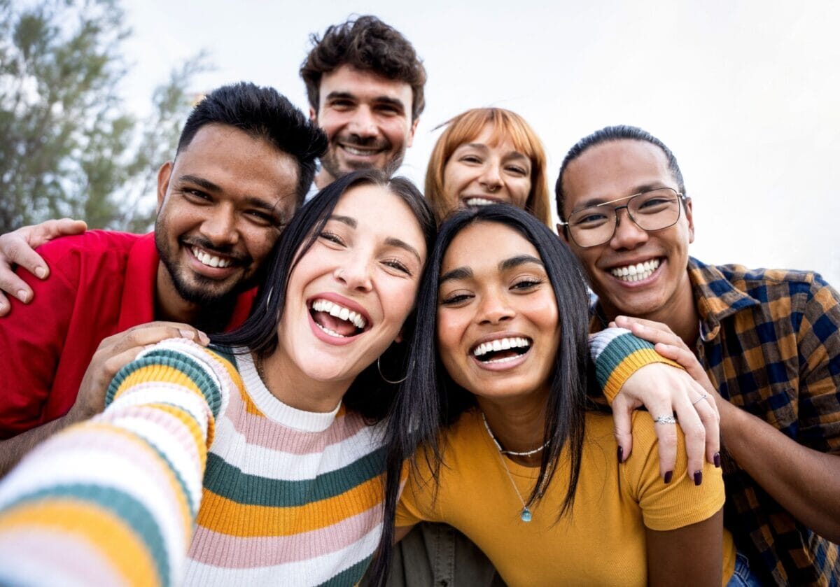 A diverse group of six friends smiling and laughing together for a close-up outdoor selfie.