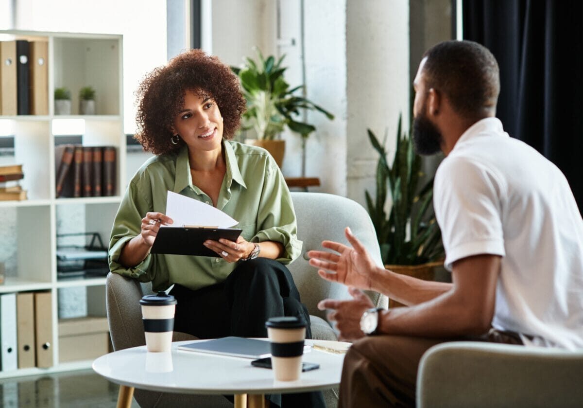 A coach smiling and holding a notebook and papers while listening attentively to a client in a bright office setting