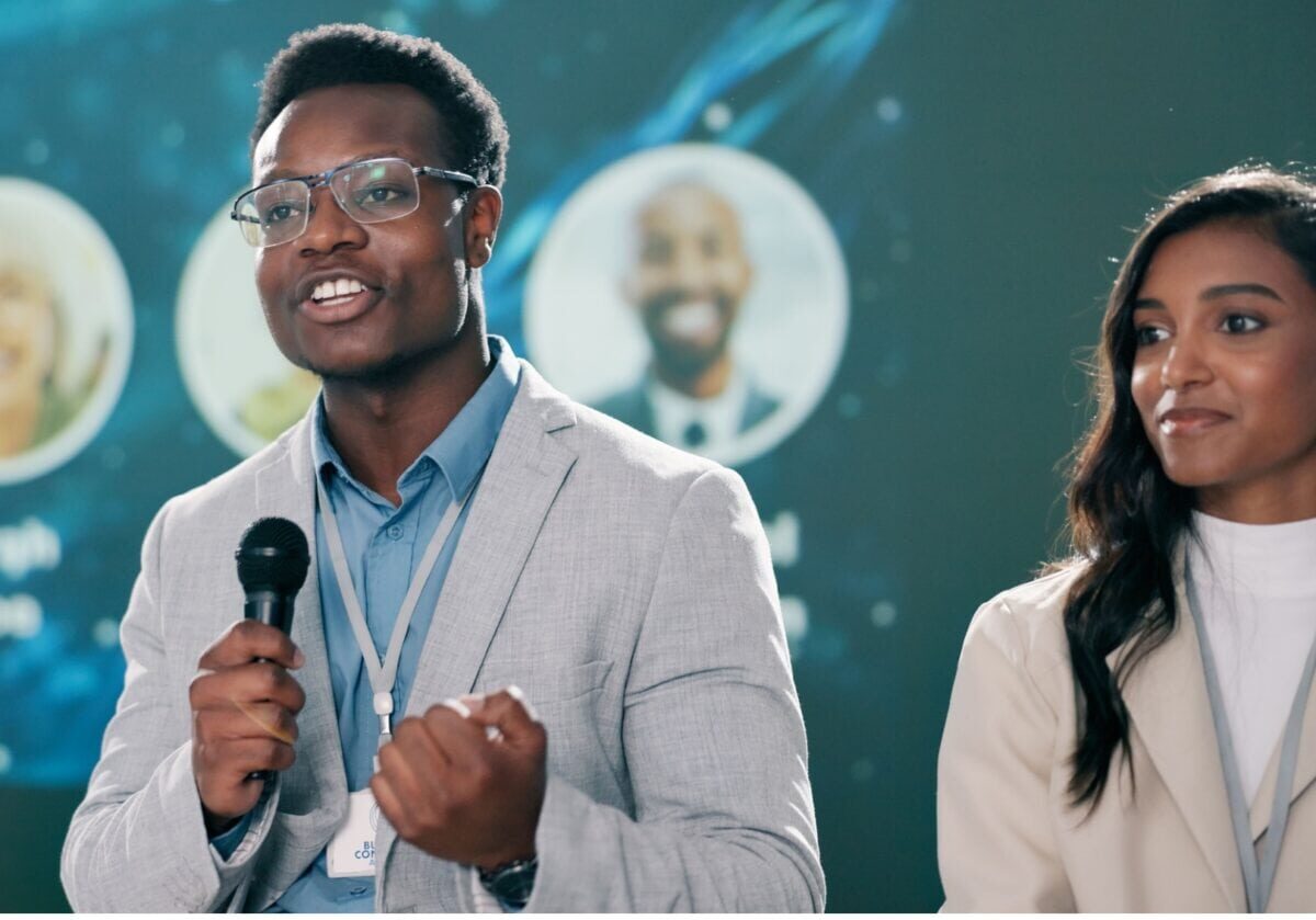 A man in a gray blazer speaks into a microphone at a professional conference while a woman beside him listens attentively.