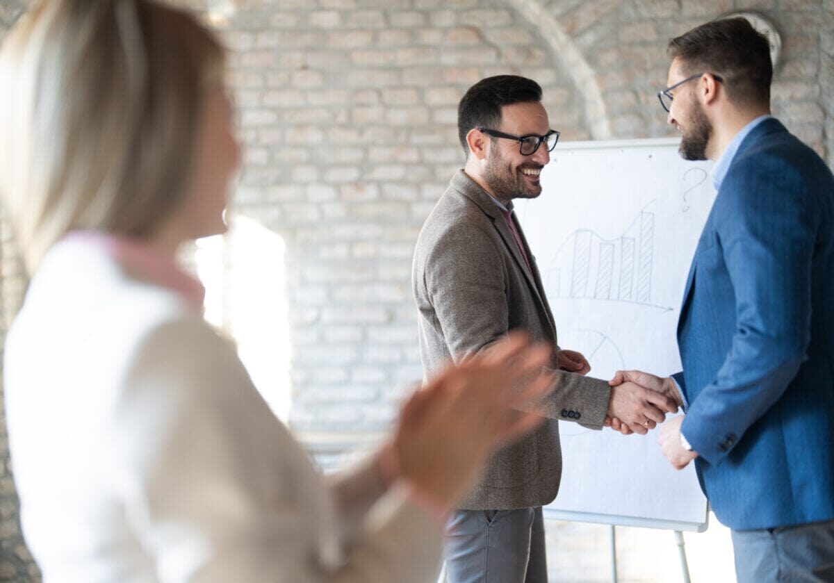 Two professionals shaking hands in front of a whiteboard chart while a colleague applauds, symbolizing career coaching and professional achievement.