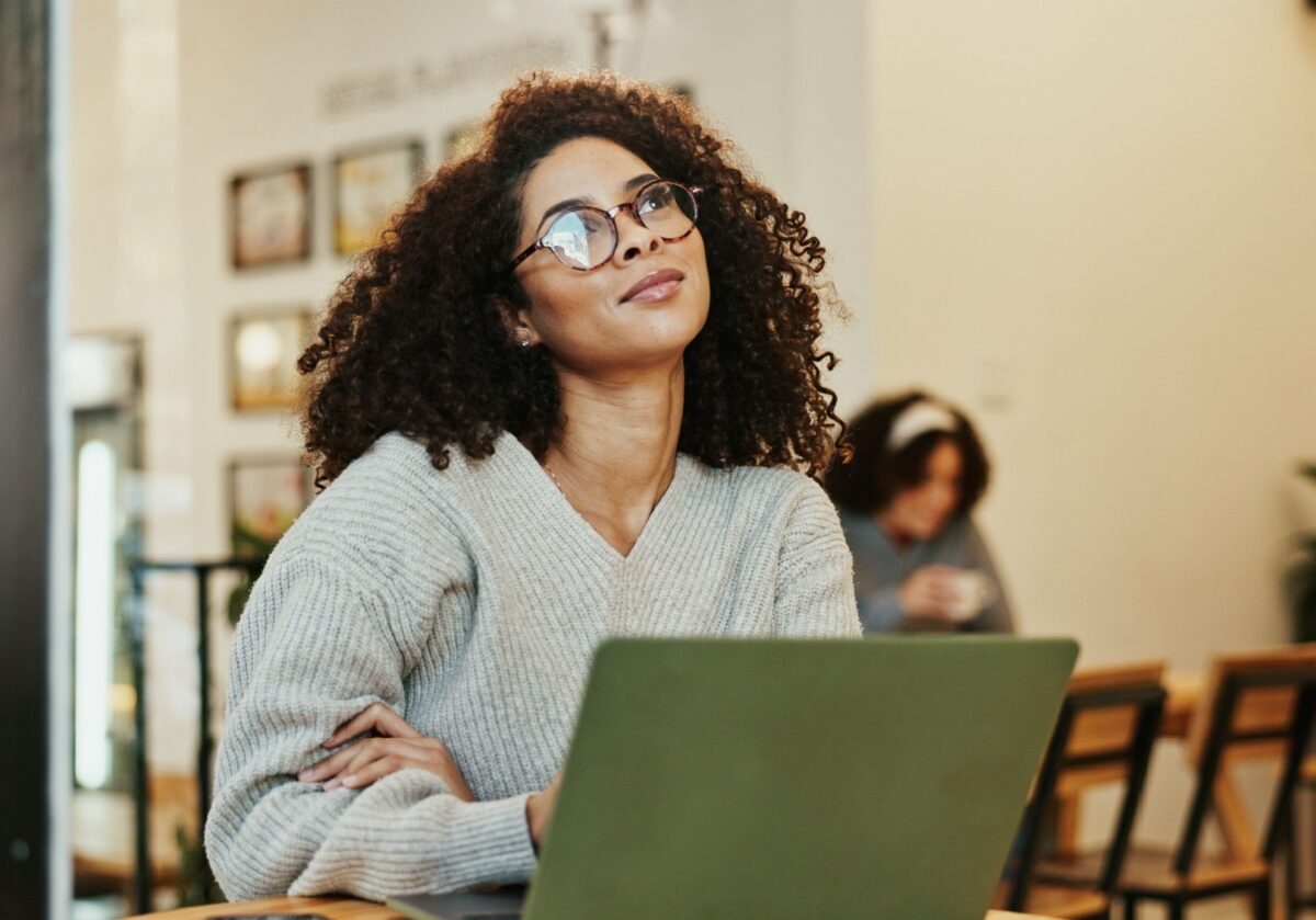 A woman with curly hair wearing glasses and a grey sweater looking up thoughtfully while sitting with her laptop in a cafe.