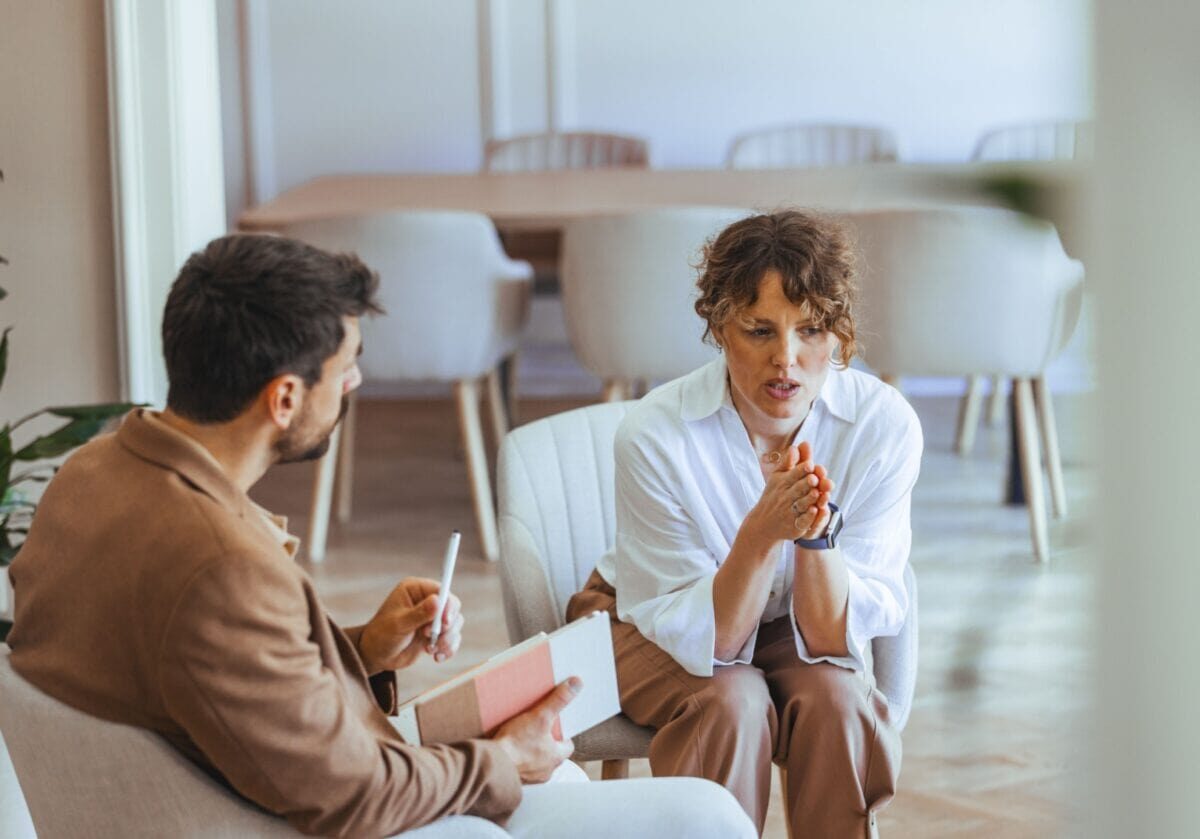 A professional woman speaks earnestly during a leadership coaching session while the coach takes notes.