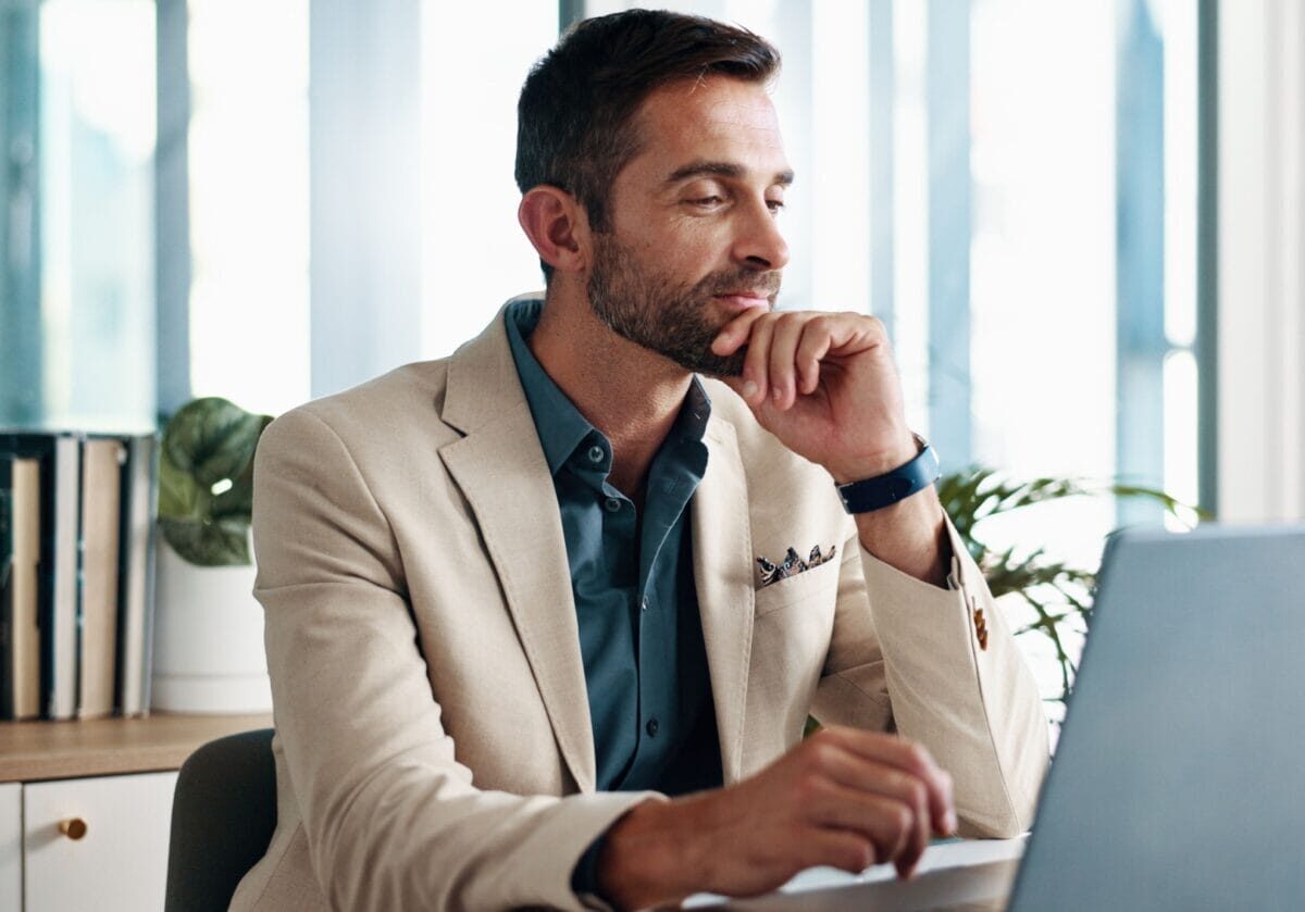 A professionally dressed man sits at a desk, gazing thoughtfully at a laptop while resting his chin on his hand.