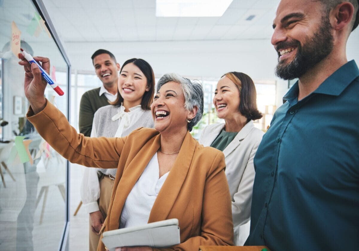 Diverse group of professionals collaborating and writing ideas on a glass board during a strategic planning discussion.