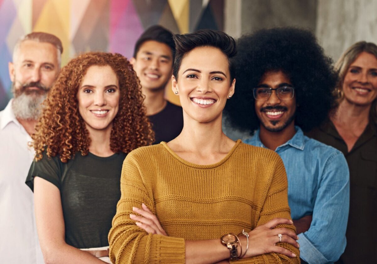 A diverse group of six smiling professionals standing together in a modern office setting, with a woman in a yellow sweater leading in the foreground.