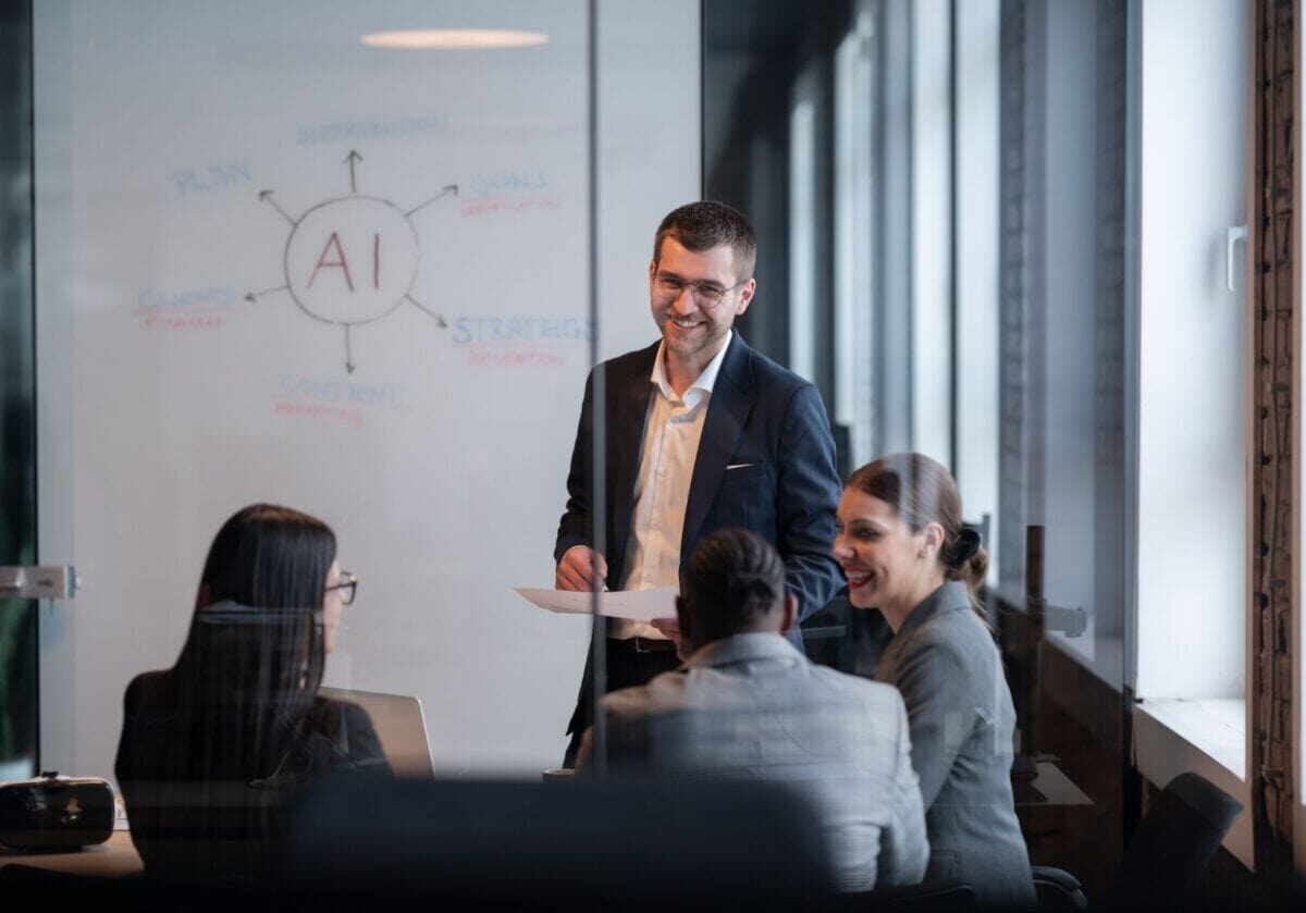 Business professionals collaborate in a meeting while discussing AI strategy, with a whiteboard displaying an AI planning diagram in the background.