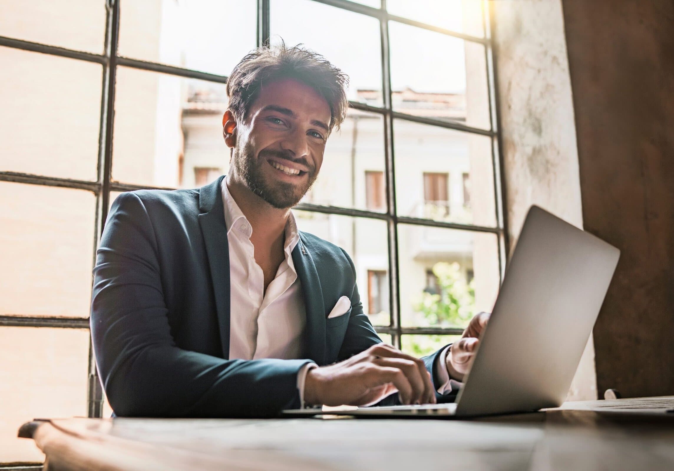 Smiling,Young,Businessman,Working,At,Laptop,Near,Window,In,Office.