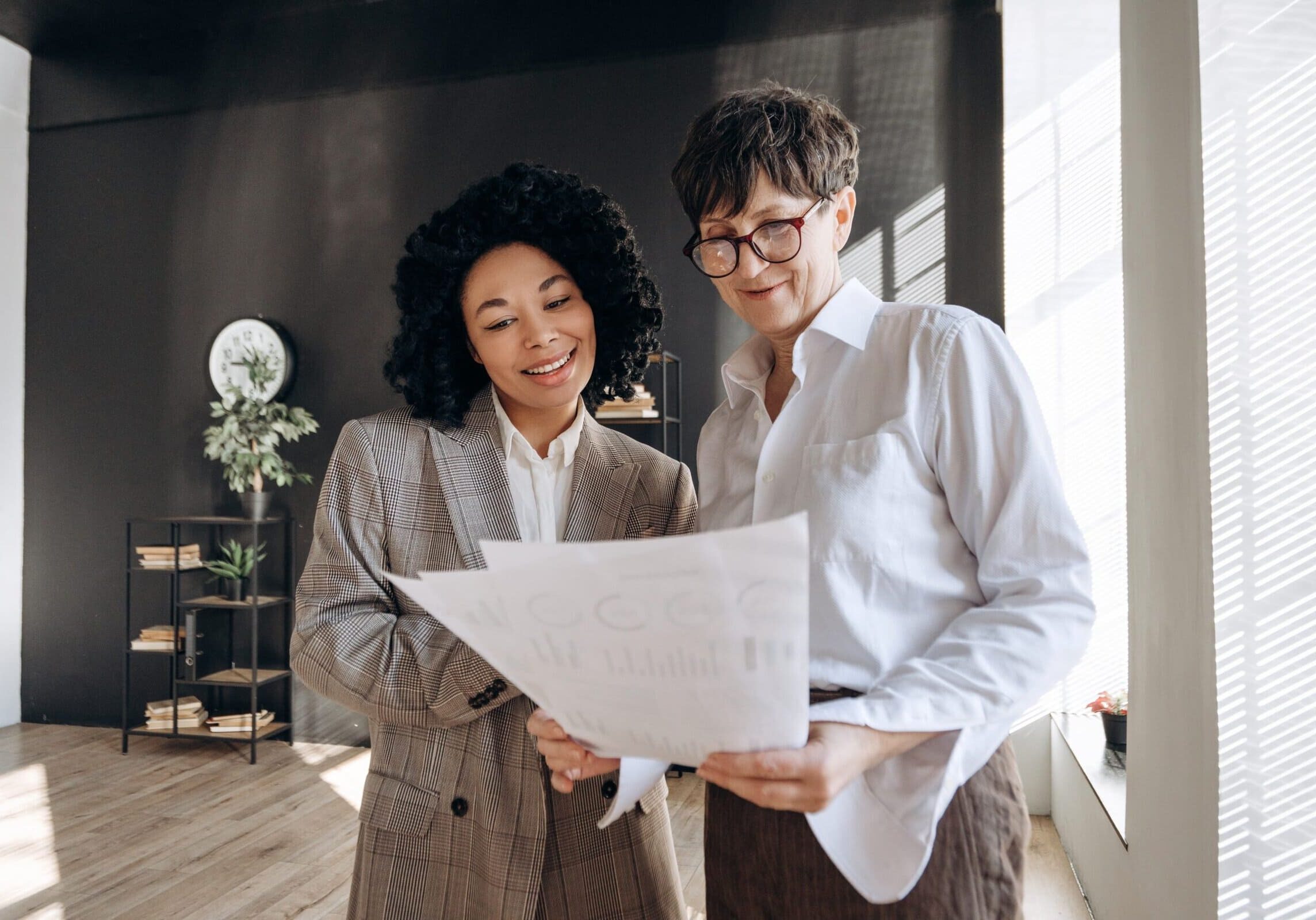 Two,Businesswomen,Smiling,And,Holding,Paperwork,With,Statistics,,Working,Together